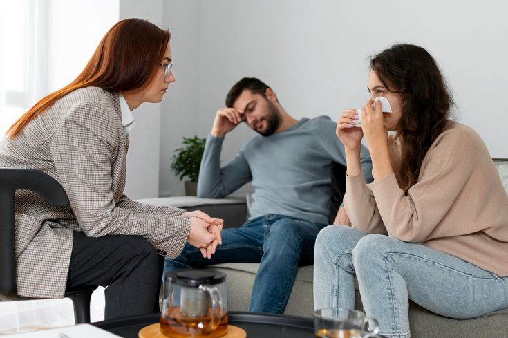 A photograph of a counseling or therapy session. On the left, a female therapist with long reddish hair and glasses wearing a plaid blazer sits in a chair, leaning forward attentively with clasped hands. On the right, a young woman sits on a grey sofa, visibly upset and holding a white tissue to her face. Sitting beside her in the background is a young man, looking distressed and resting his head against his hand. A black coffee table with a glass teapot and cups sits in the foreground.