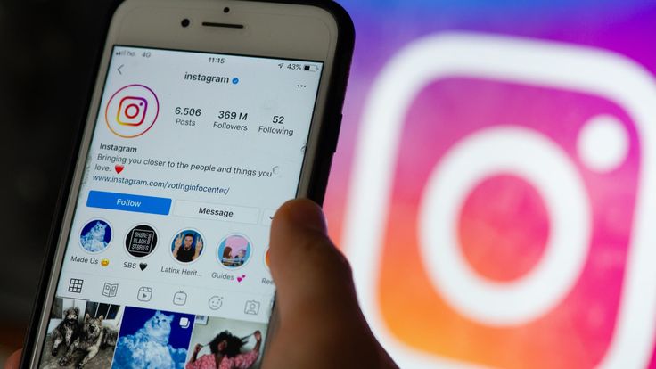A close-up of a hand holding a white smartphone, displaying the official Instagram profile page on the screen. The profile shows 369 million followers, a blue 'Follow' button, story highlights, and a grid of recent posts. In the blurred background, a large, vibrantly lit Instagram logo is displayed on a computer monitor.