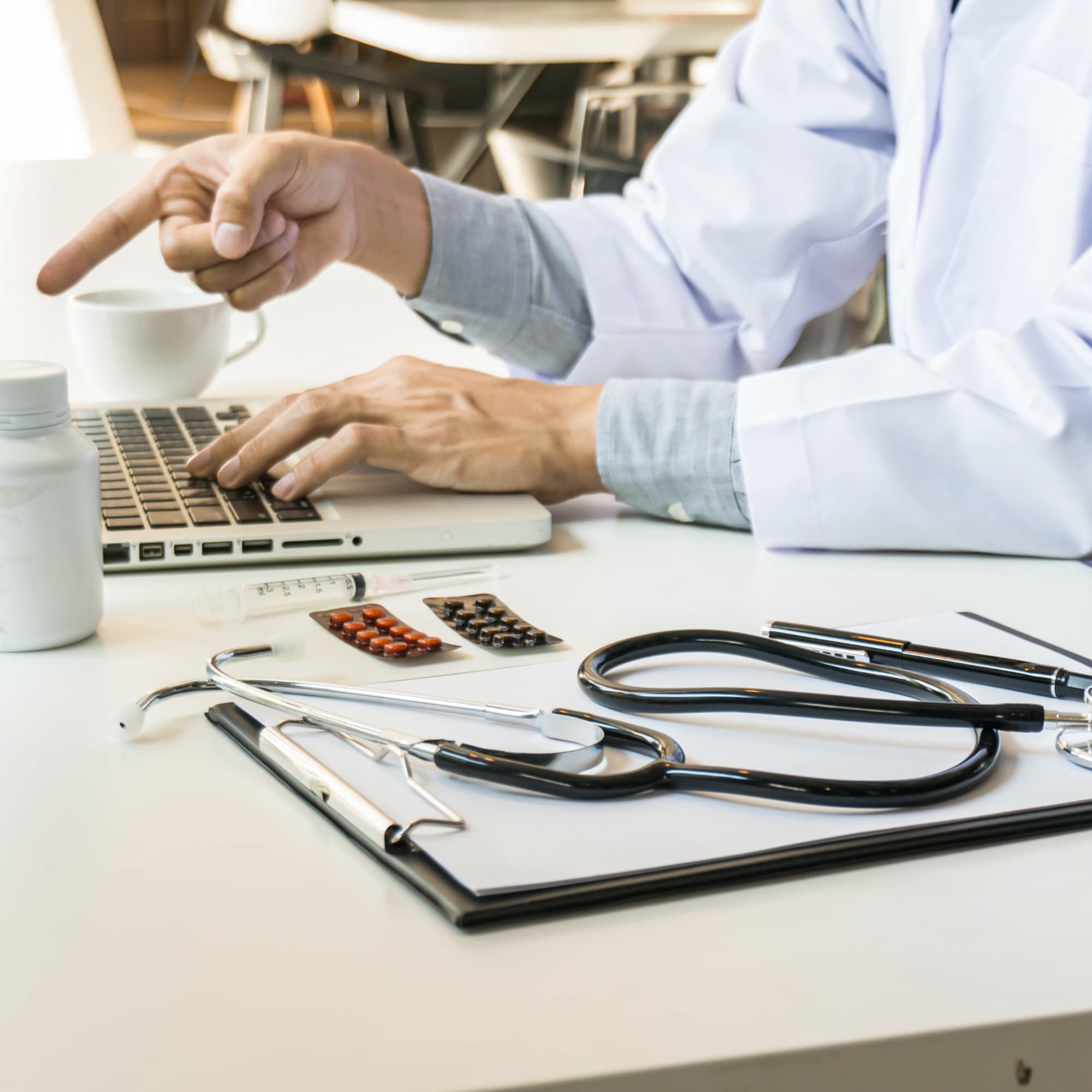 A doctor working on a laptop at a desk with medical tools, including a stethoscope, syringe, pills, and clipboard, suggesting a clinical workspace or telehealth setup.