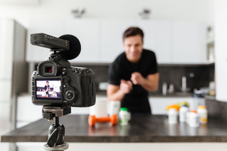 A high-quality photo of a professional video production setup in a modern kitchen. A DSLR camera with a top-mounted microphone sits on a tripod in the foreground, with its digital screen displaying a live shot of a male creator. The creator is visible in the background, slightly out of focus, gesturing enthusiastically while recording a video next to a display of fitness supplement bottles.