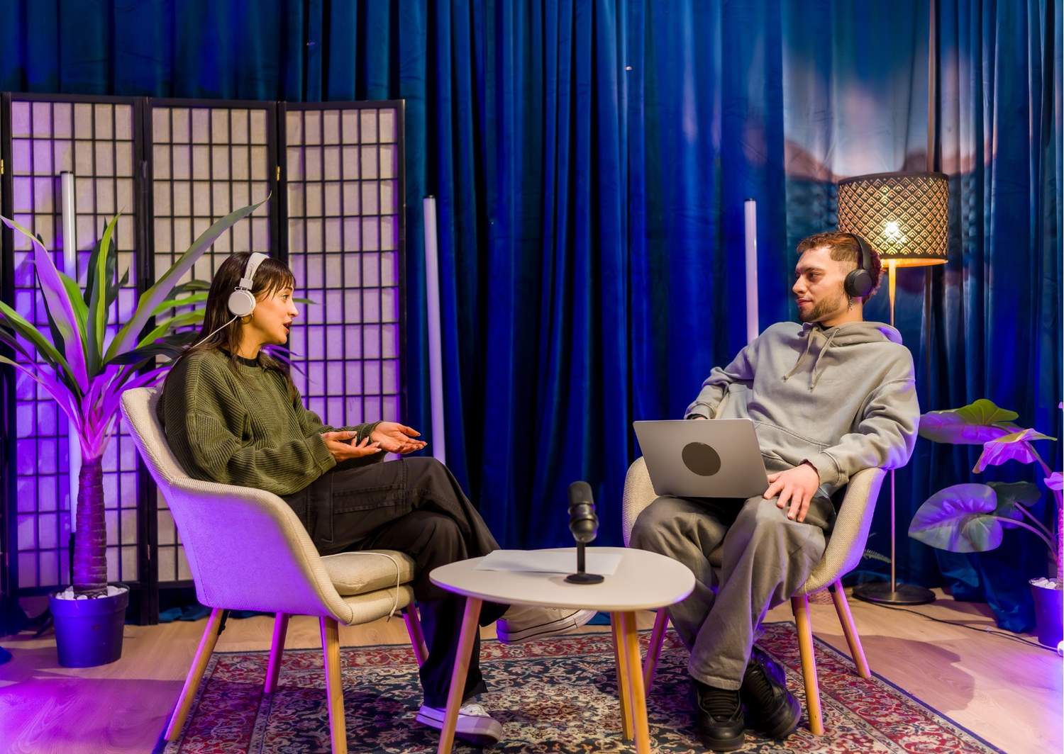  Gemini said Two podcasters, a woman and a man, are engaged in a professional recording session within a stylized studio. Both wear headphones and are seated in comfortable armchairs facing each other across a small coffee table equipped with a microphone.