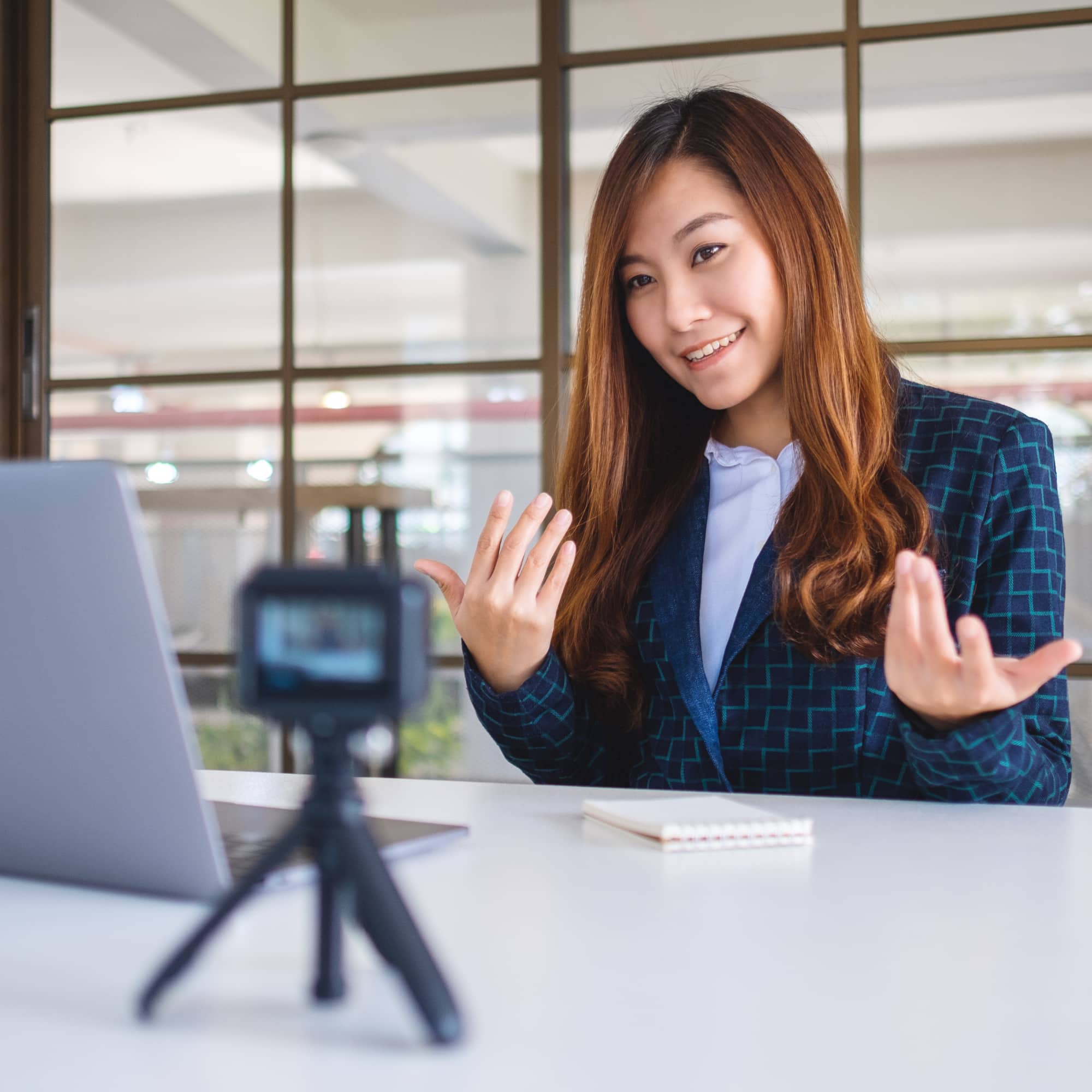 Woman in a blazer speaking and gesturing while recording a video on a camera, sitting at a desk with a laptop and notebook.