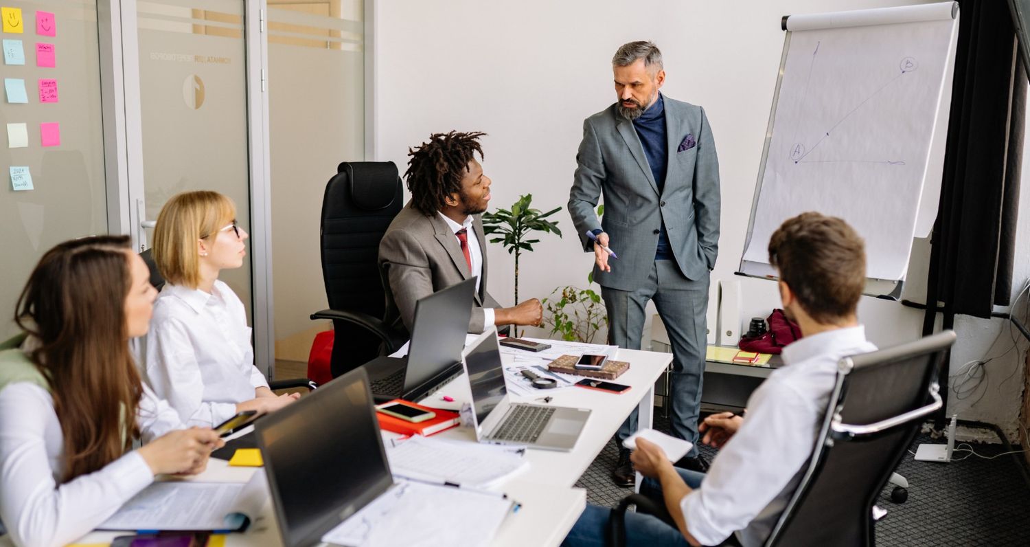 A business coach in a grey suit presents a graph on a whiteboard to a diverse group of four young professionals at a conference table in a modern office.