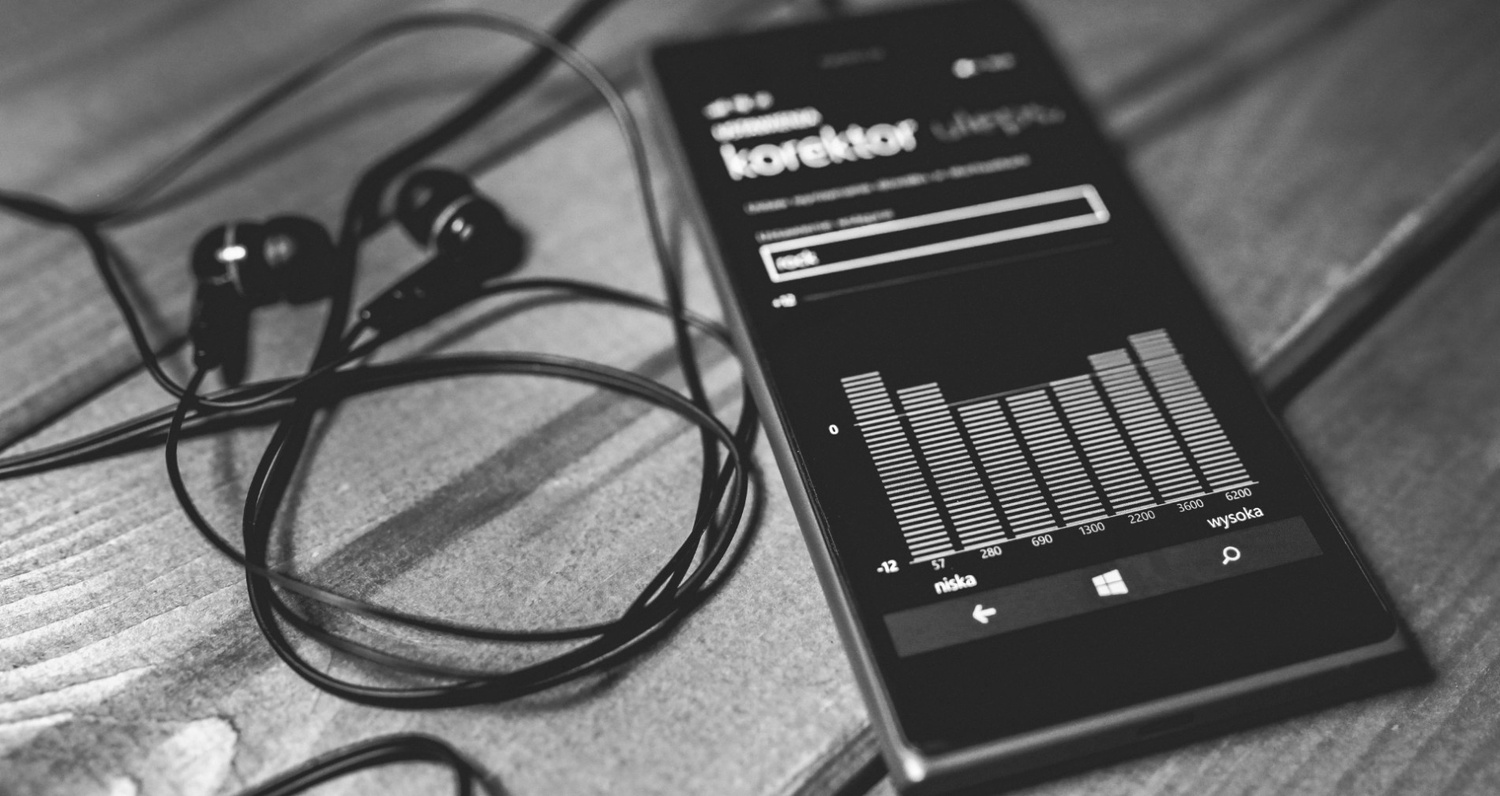 Black and white close-up of a smartphone displaying an audio equalizer app next to a pair of wired earphones on a wooden surface.