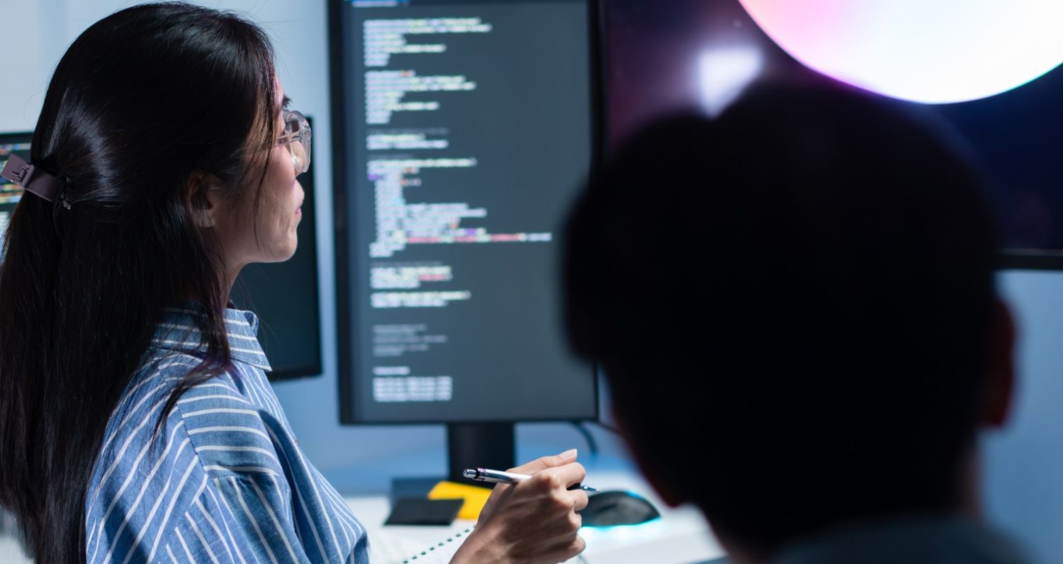 Gemini said
A woman with glasses and a striped shirt looking at a computer screen filled with lines of code while holding a pen, in a dark office setting with a colleague in the foreground.