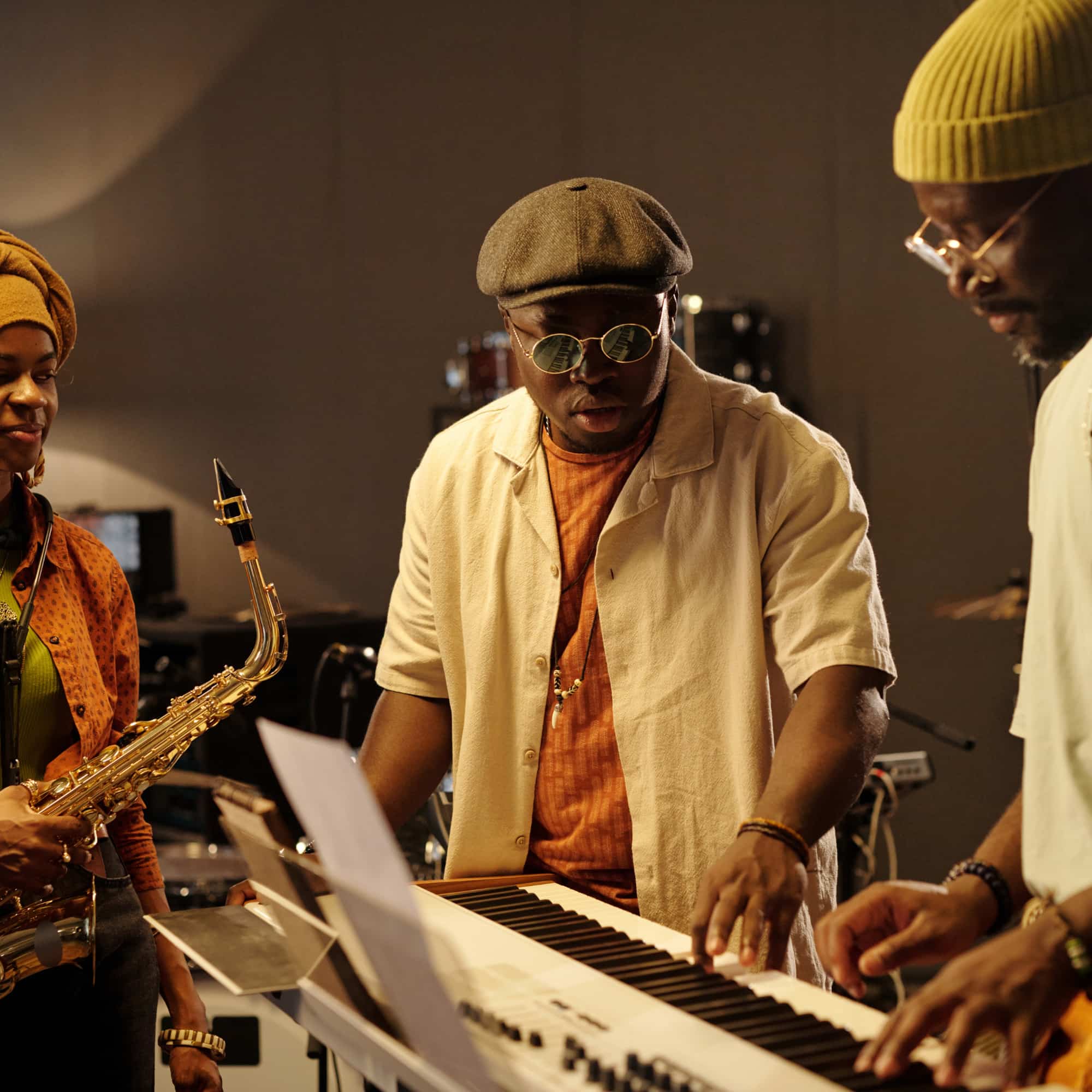 Three musicians rehearsing in a studio, with one playing a keyboard while another holds a saxophone and a third adjusts controls, focused on their performance.