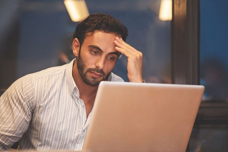 A stressed man resting his hand on his forehead while looking at his laptop screen.