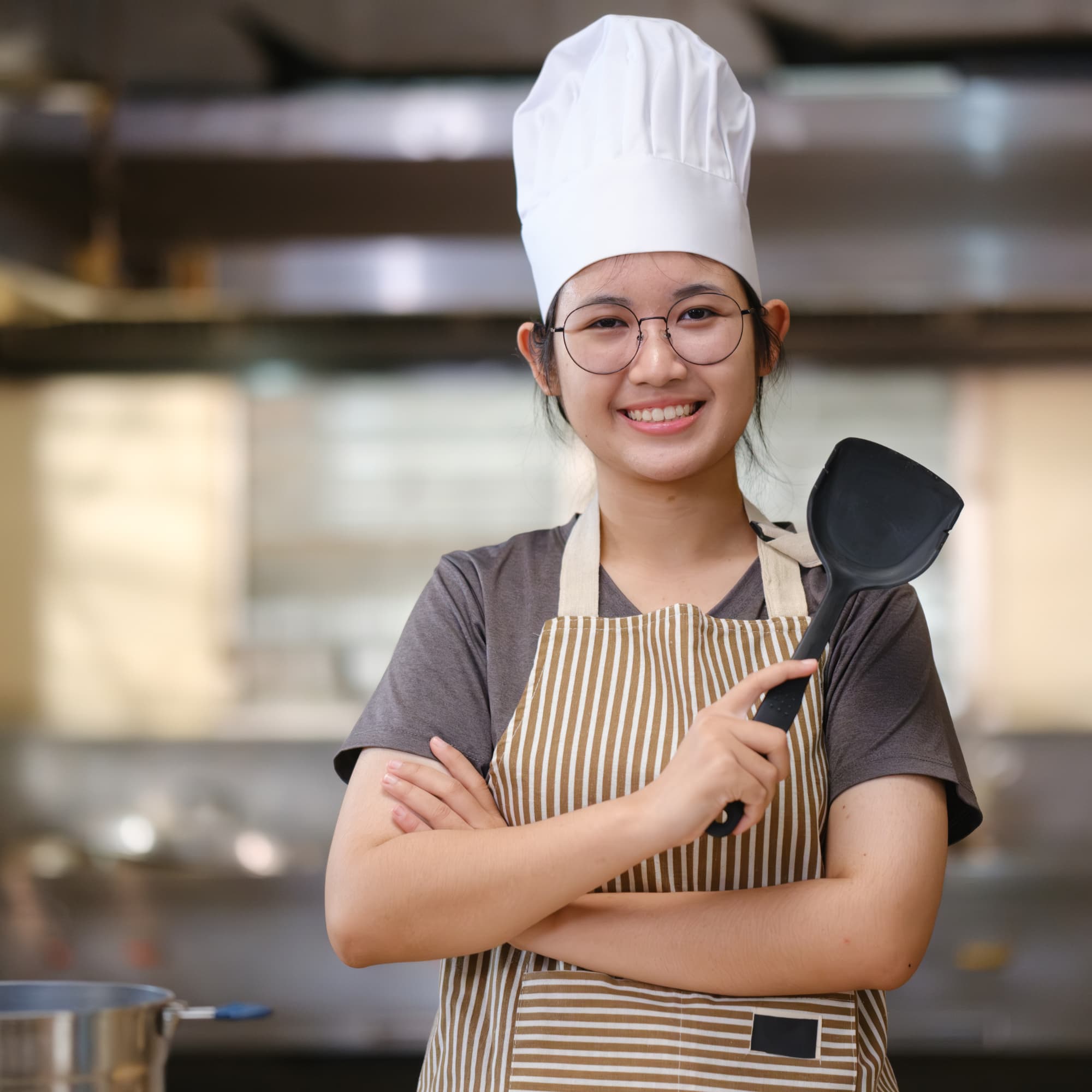 Smiling chef wearing a hat and apron, holding a spatula in a professional kitchen.