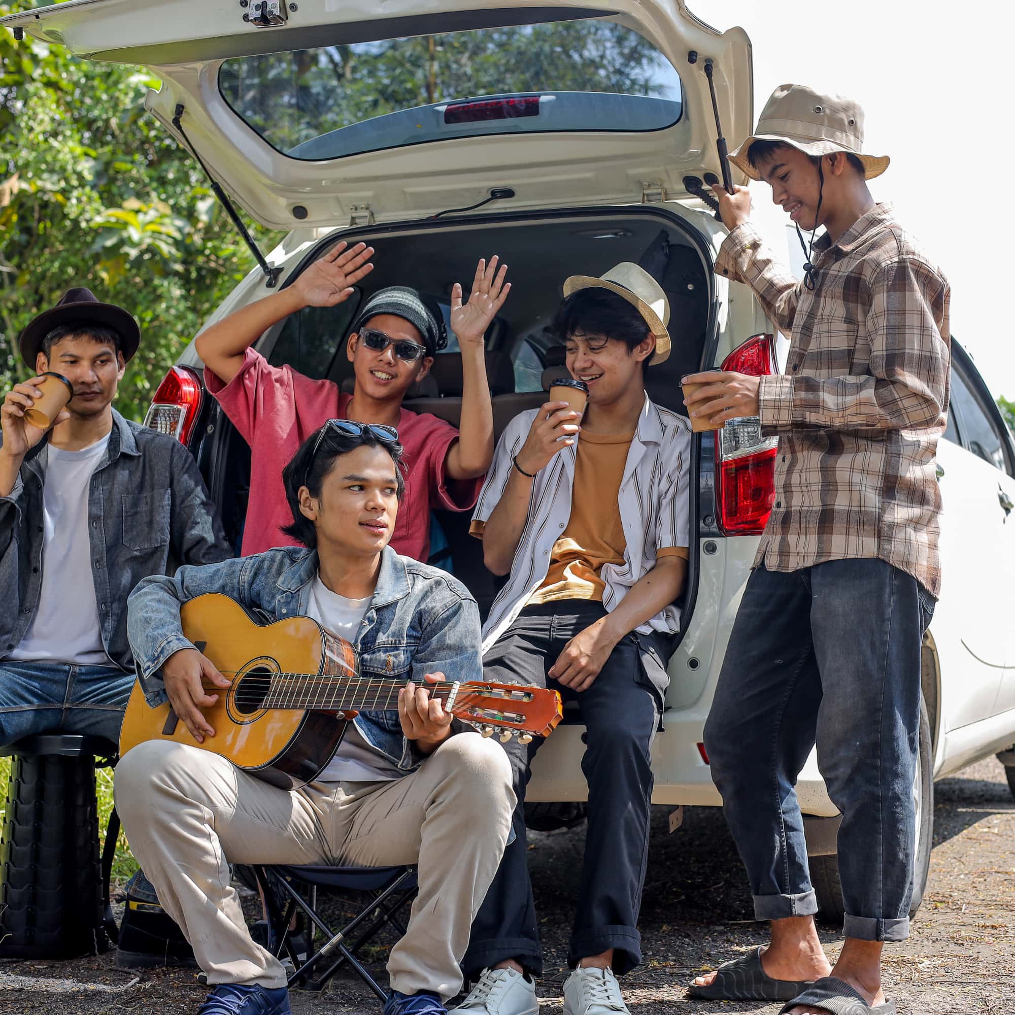 Group of friends relaxing outdoors by an open car trunk, with one playing guitar while others drink coffee and chat in a casual setting.