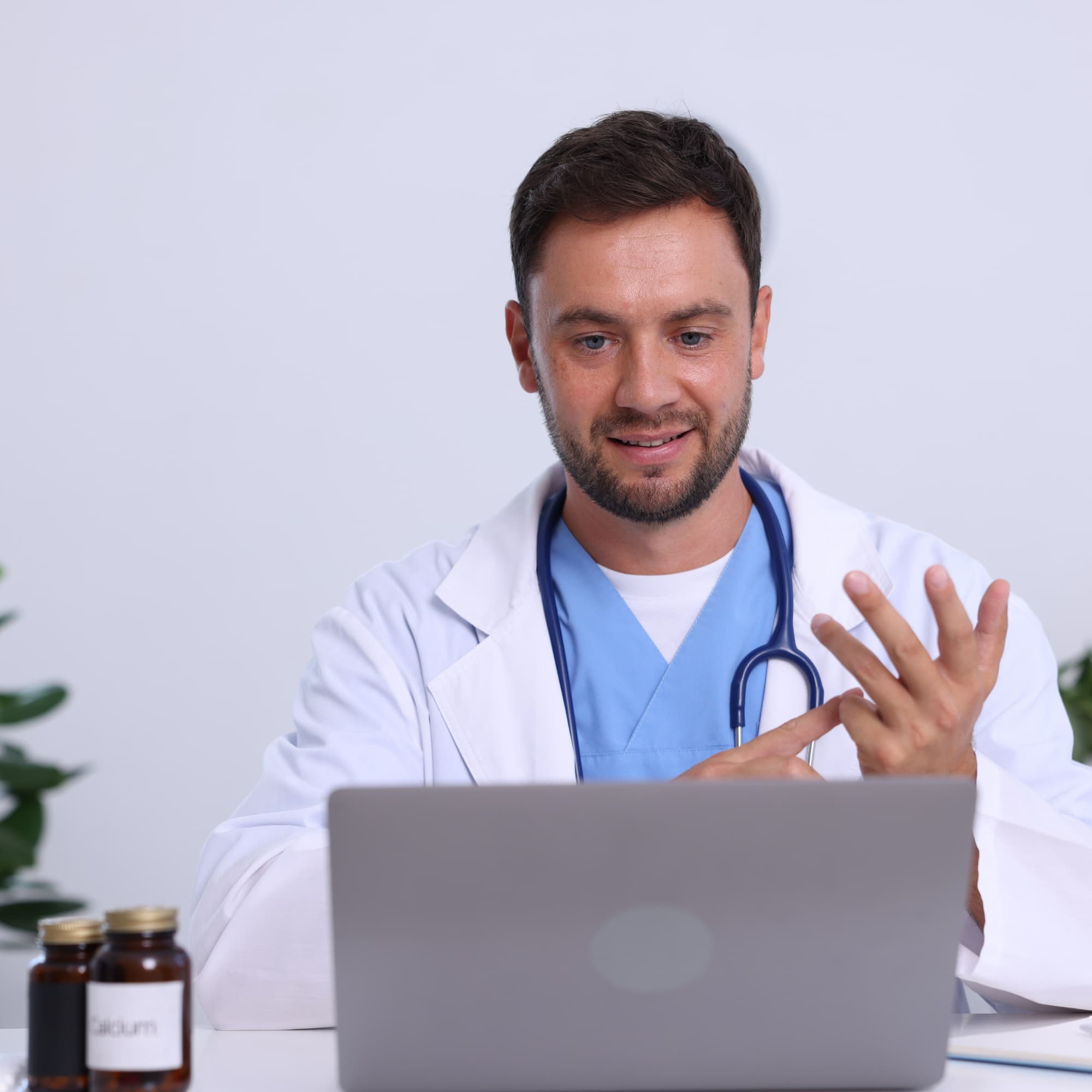 A doctor in a white coat and stethoscope using a laptop while gesturing with fingers, as if explaining something during an online consultation.