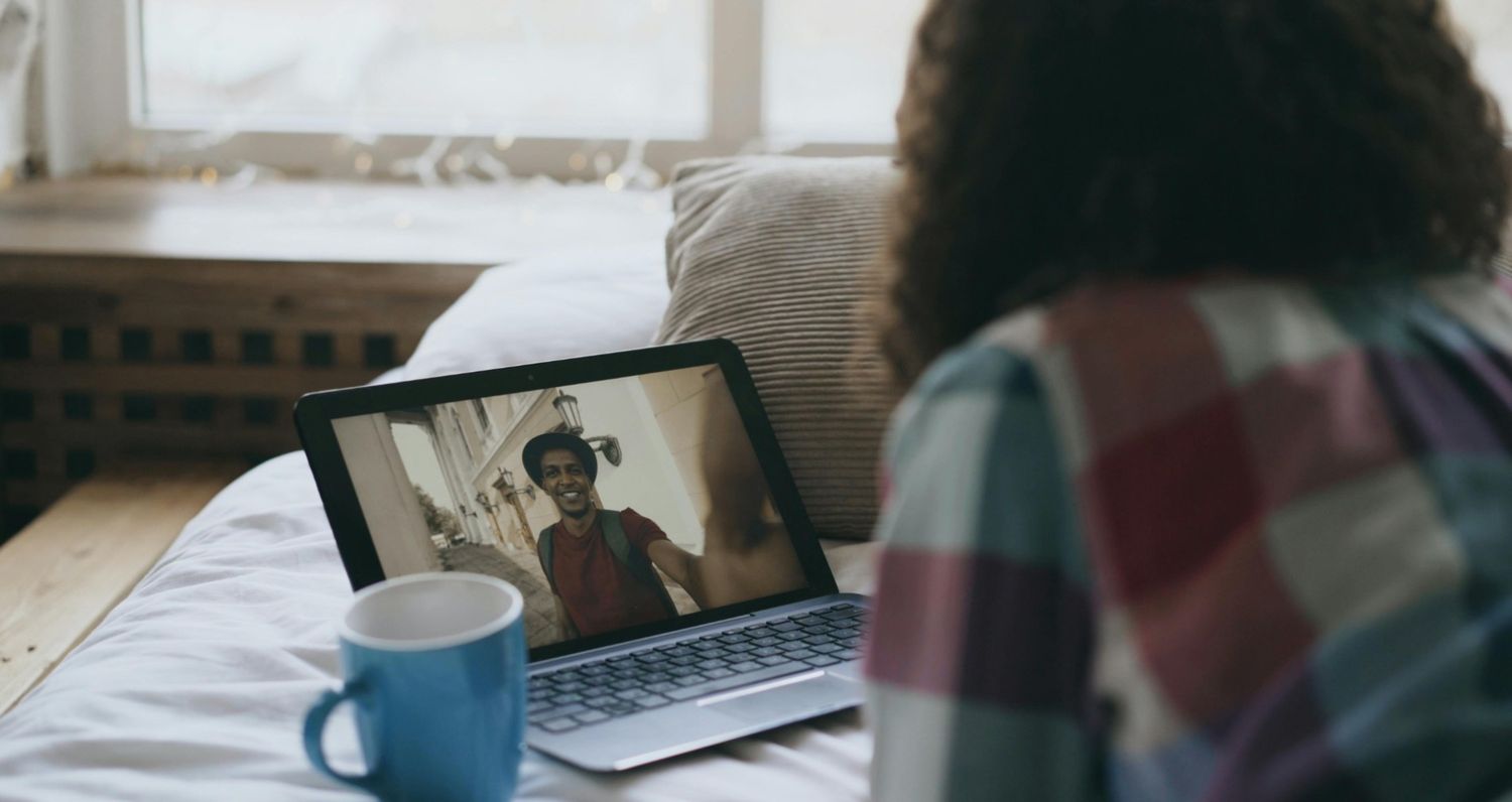 Over-the-shoulder view of a person sitting on a bed with a blue mug, watching a video on a laptop that shows a man in a hat smiling and recording a selfie-style vlog outdoors.