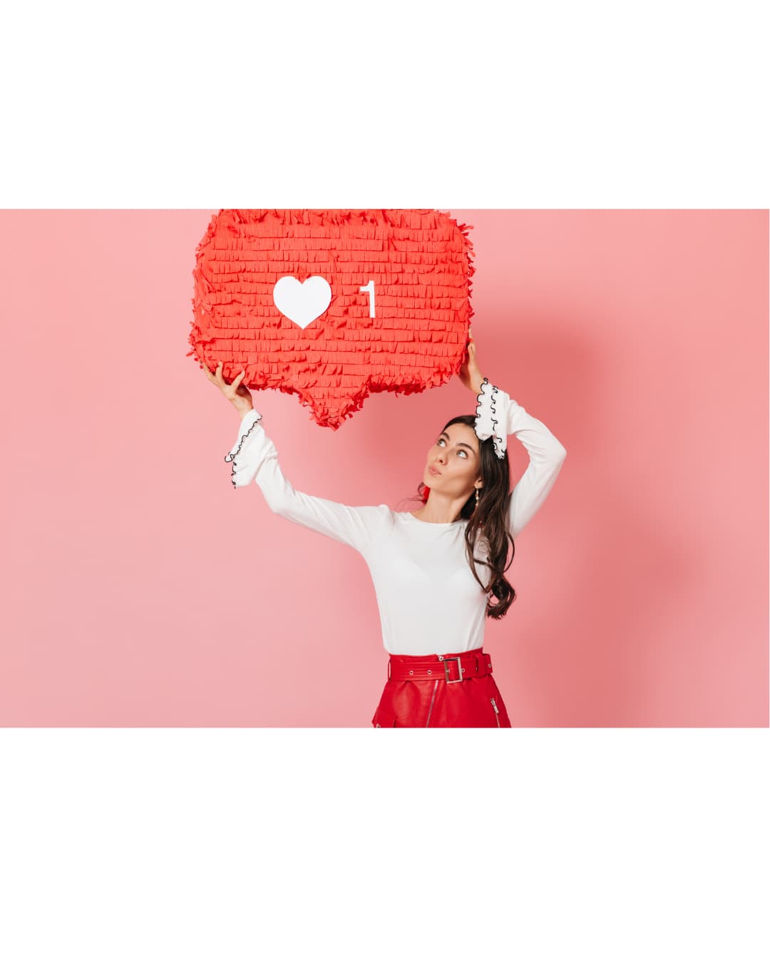 A woman stands against a pink background holding a large social media like notification prop above her head. The oversized red icon features a white heart and the number '1,' symbolizing likes, social media engagement, online interactions, influencer marketing, and digital popularity.