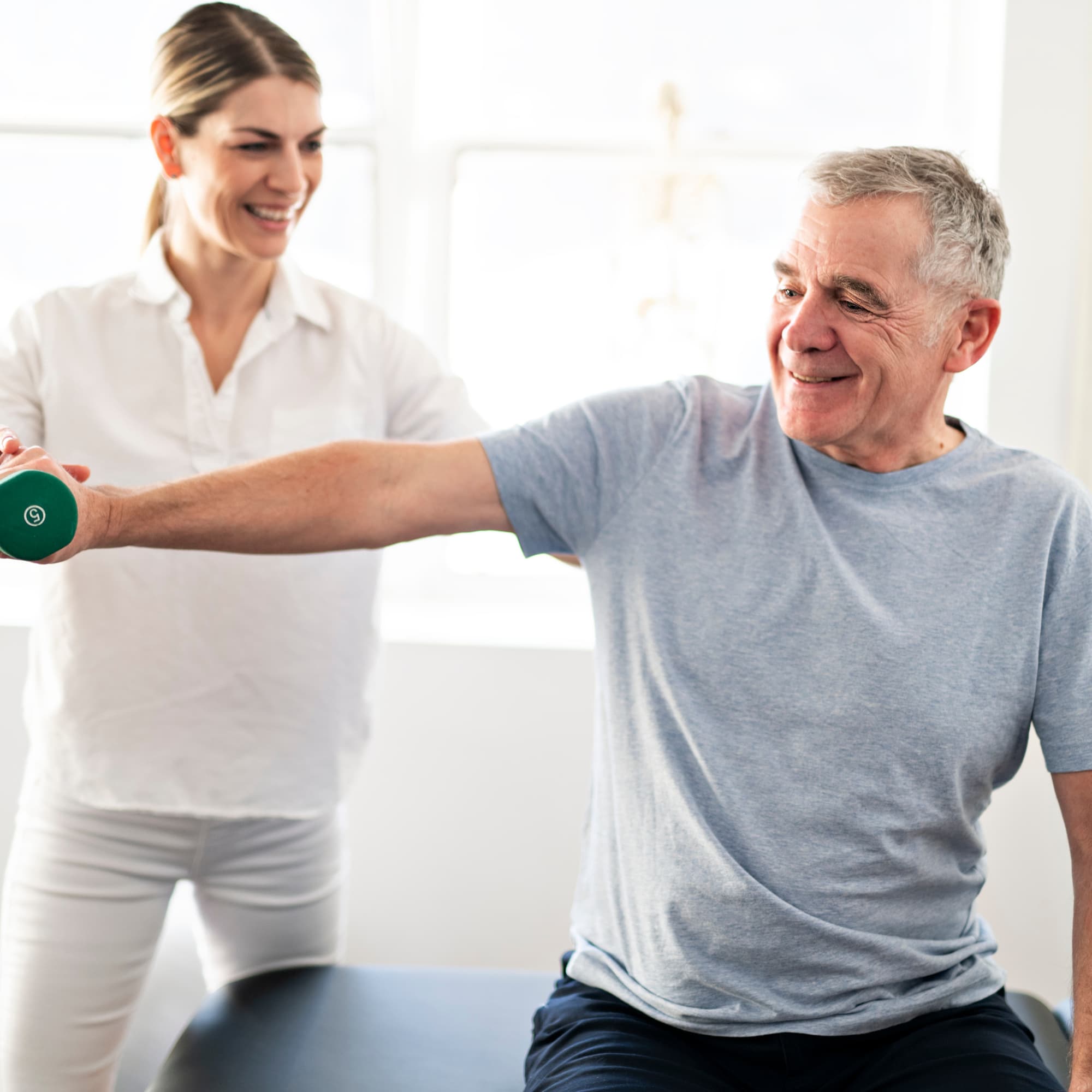 A physiotherapist guiding a senior man through a light dumbbell exercise, supporting strength training and rehabilitation in a bright clinical environment.