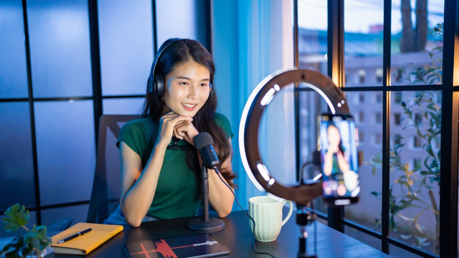 A smiling young woman wearing headphones, speaking into a professional microphone while recording a video on a smartphone mounted inside a ring light.