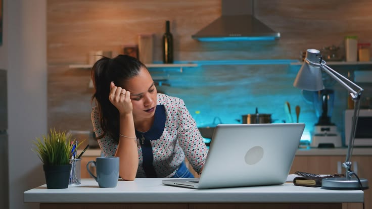A tired young woman leaning her head on her hand while staring at a laptop screen late at night in a kitchen setting. She appears exhausted or burnt out, symbolizing the struggle of choosing quantity over quality in content creation. The room is dimly lit with blue accent lighting in the background and a warm desk lamp in the foreground.