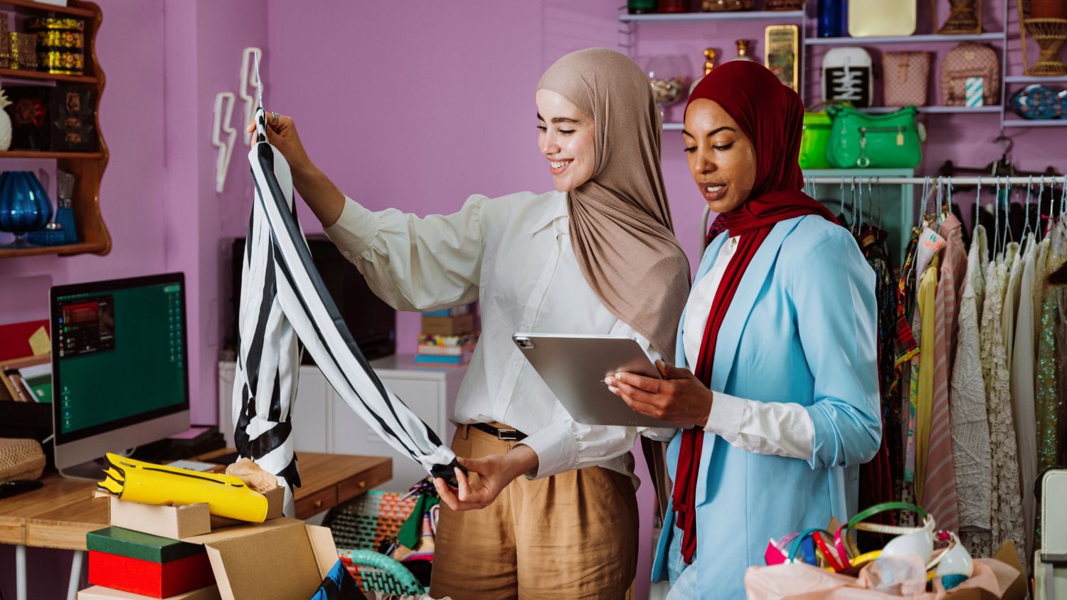 Two smiling women wearing hijabs collaborate in a colorful clothing boutique; one holds a striped garment while the other manages inventory on a digital tablet.