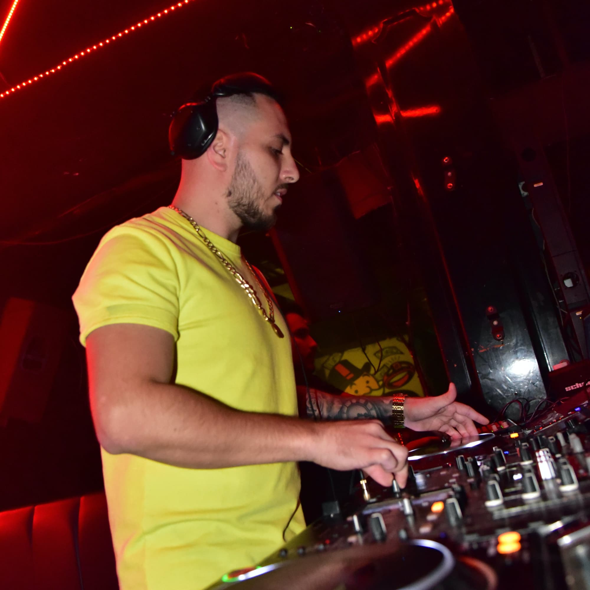 Male DJ wearing headphones and a yellow shirt, focused on mixing music on a DJ controller in a dimly lit club with red lighting.