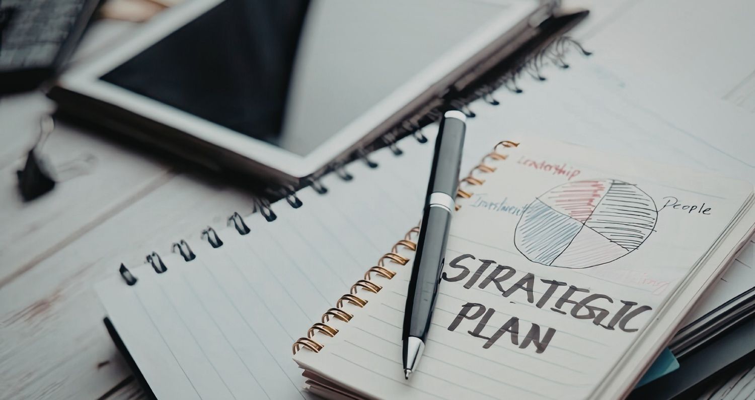 A close-up shot of a spiral notebook on a white wooden desk. A pen rests on a page featuring a hand-drawn pie chart with labels like 'Leadership,' 'People,' and 'Investment,' alongside the large handwritten words 'STRATEGIC PLAN.
