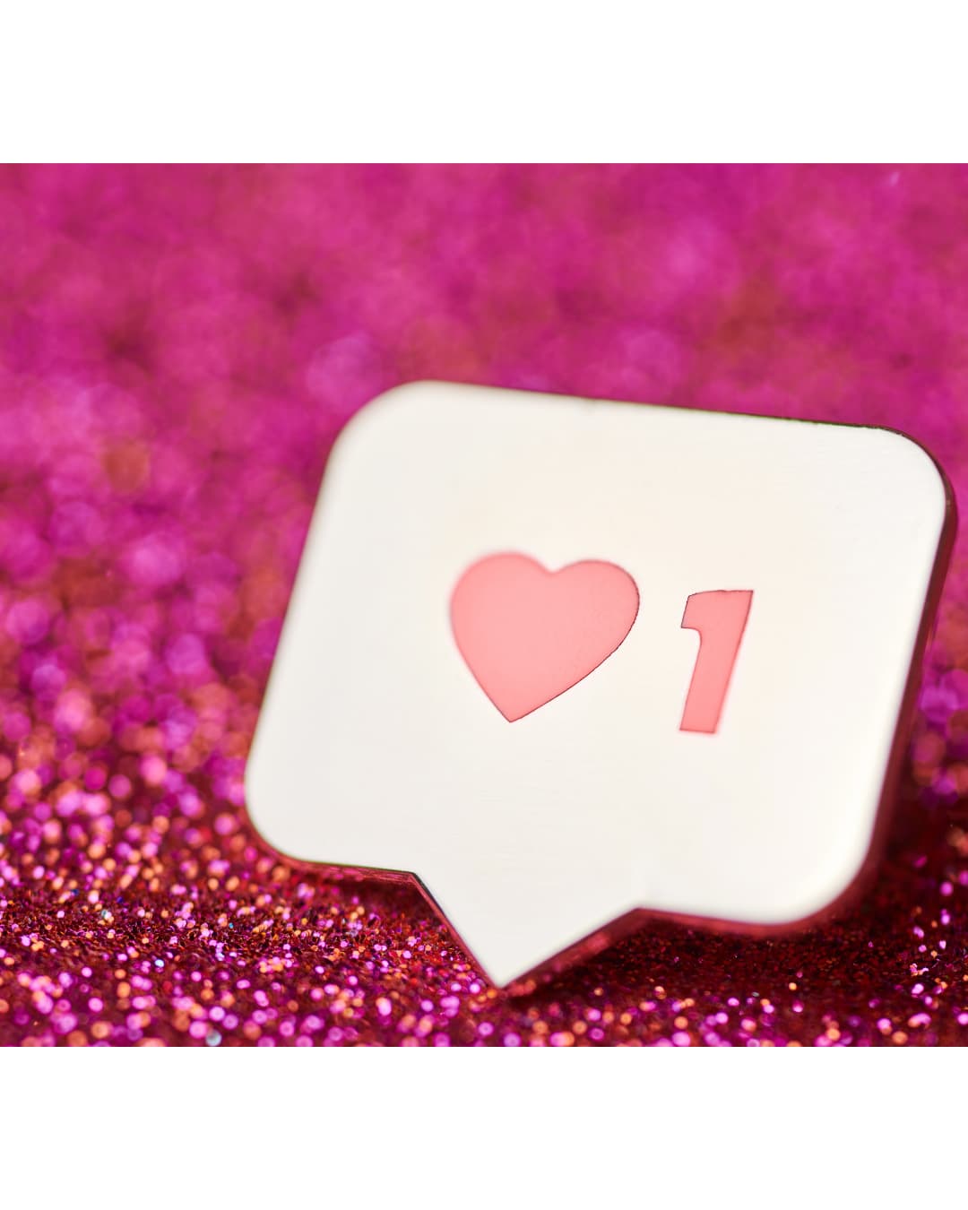A social media notification bubble with a heart icon and the number 1 on a sparkling pink glitter background.