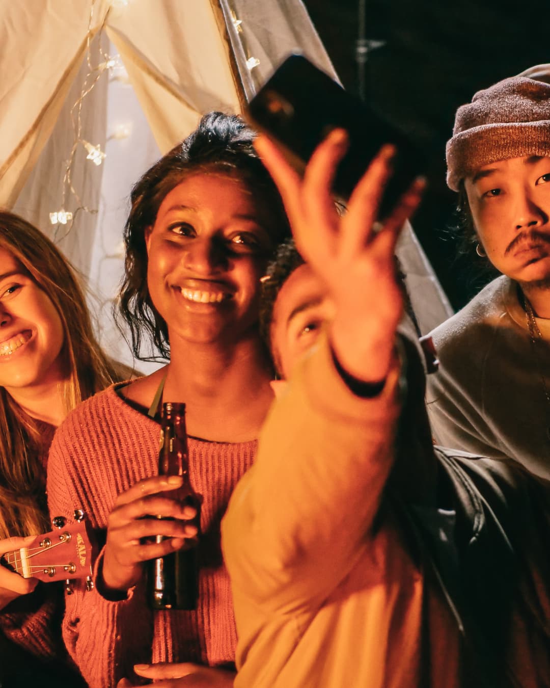 A group of friends gathers in a cozy, warmly lit setting, smiling as one person holds up a smartphone to take a selfie. String lights and casual attire create a relaxed social atmosphere, capturing a moment of friendship, celebration, and social media sharing.