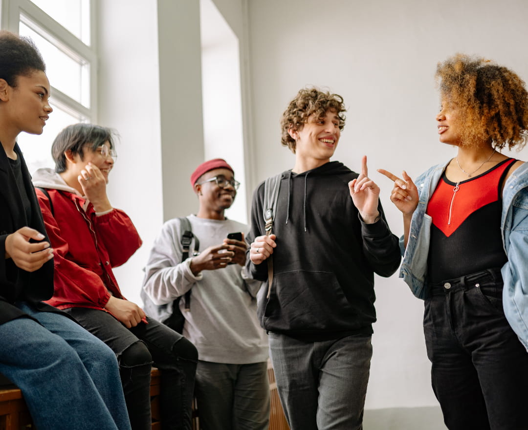 A group of five young people standing indoors near a window, casually chatting and smiling. Two individuals in the center are engaged in a lively conversation, gesturing with their hands, while the others listen and interact informally. The setting appears bright and relaxed, suggesting a friendly social or school environment.