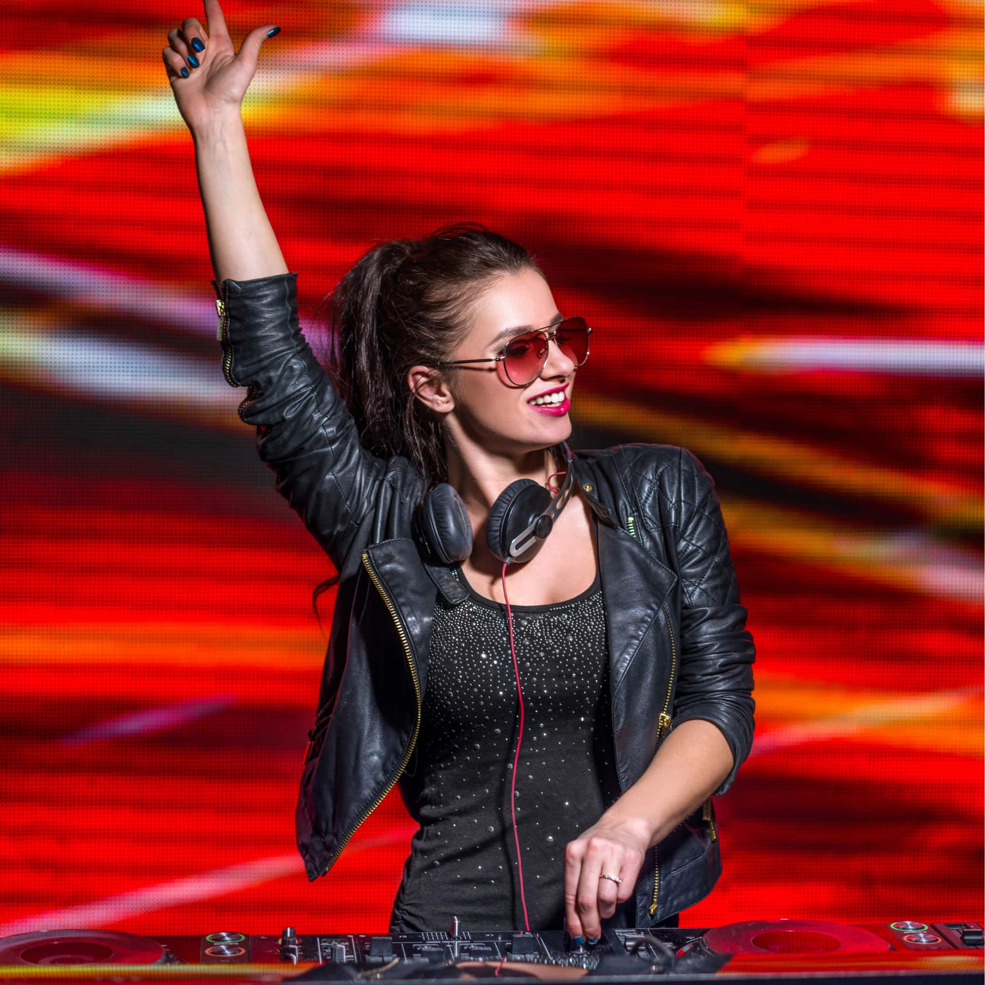 Female DJ wearing a black leather jacket and red sunglasses, smiling while raising her hand and mixing music on a DJ controller against a vibrant red light background.