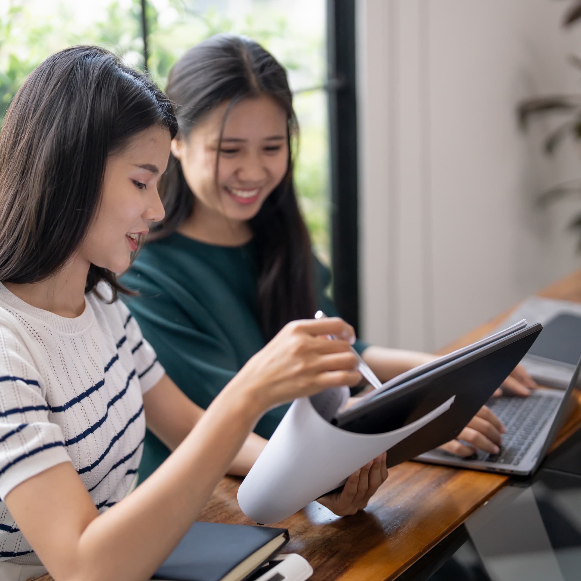 Two colleagues collaborating at a desk, reviewing documents on a clipboard while working on a laptop.