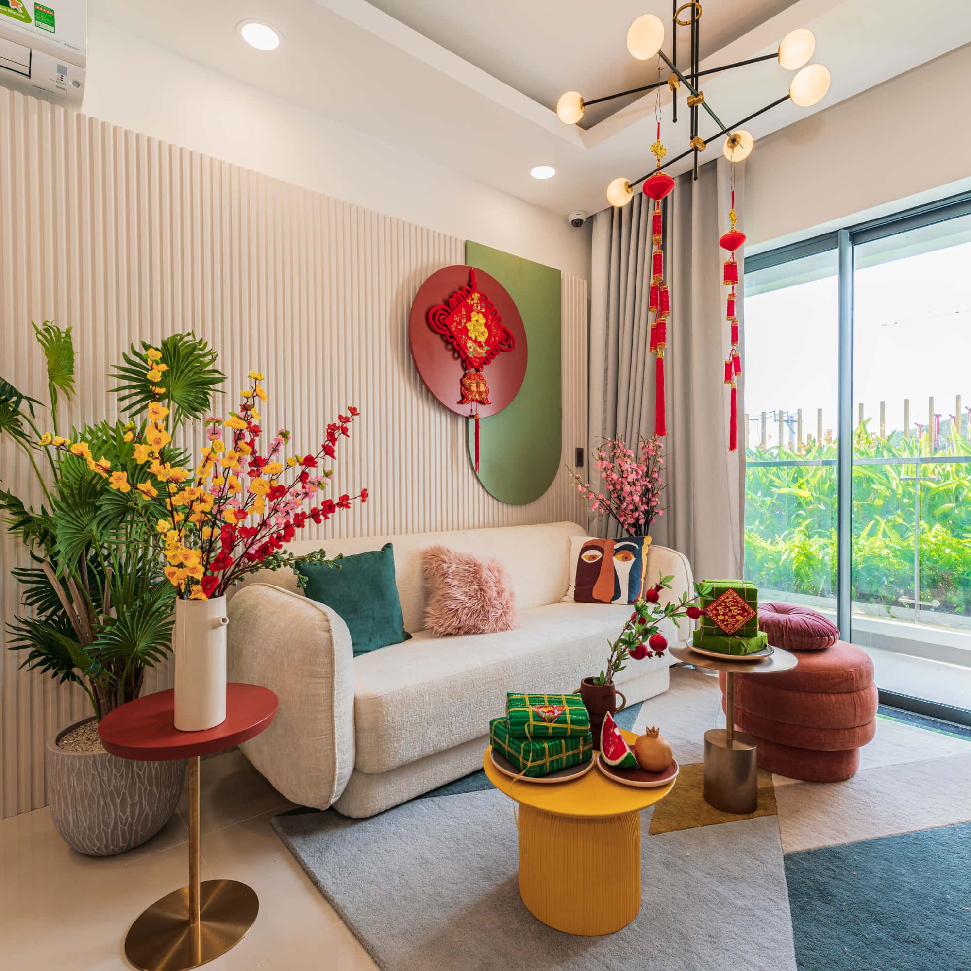 Modern living room decorated for a festive celebration with red hanging ornaments, floral arrangements, a neutral sofa, colorful cushions, and a bright balcony view.