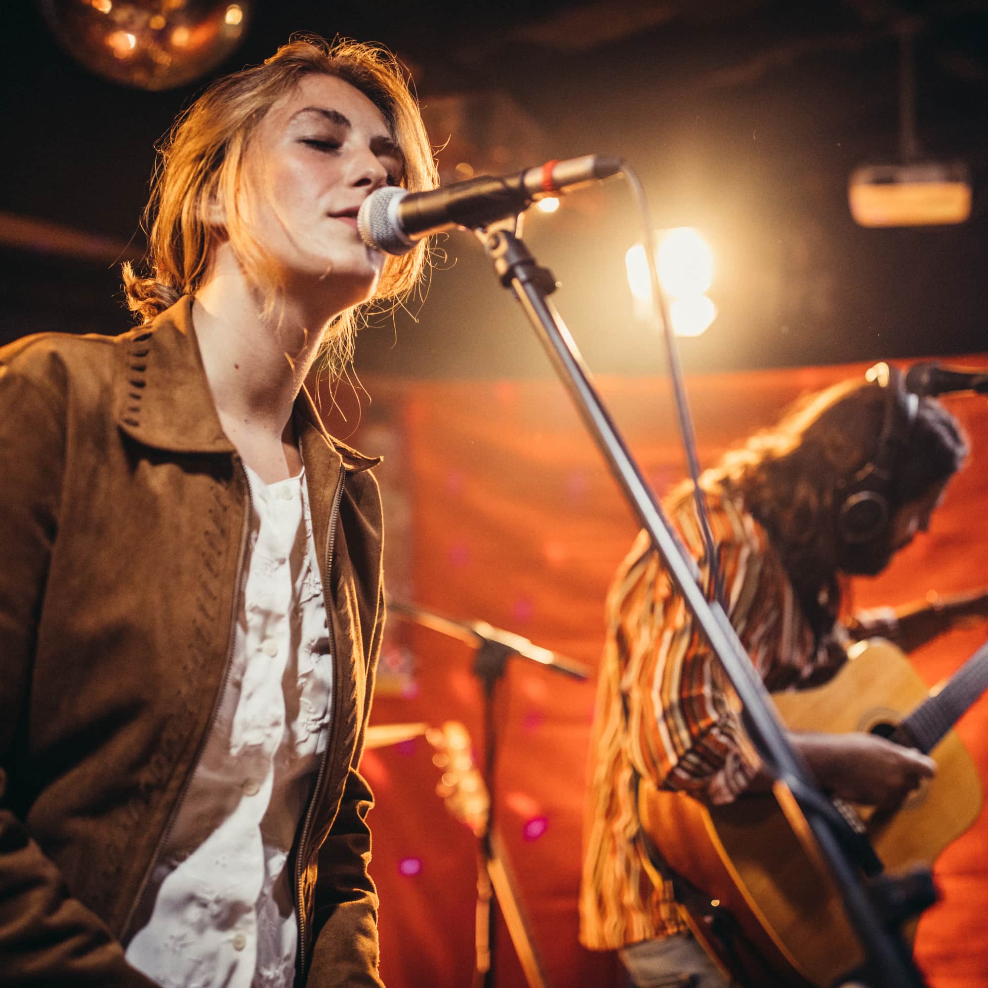 Female singer performing live on stage, singing into a microphone while a guitarist plays in the background under warm lighting.
