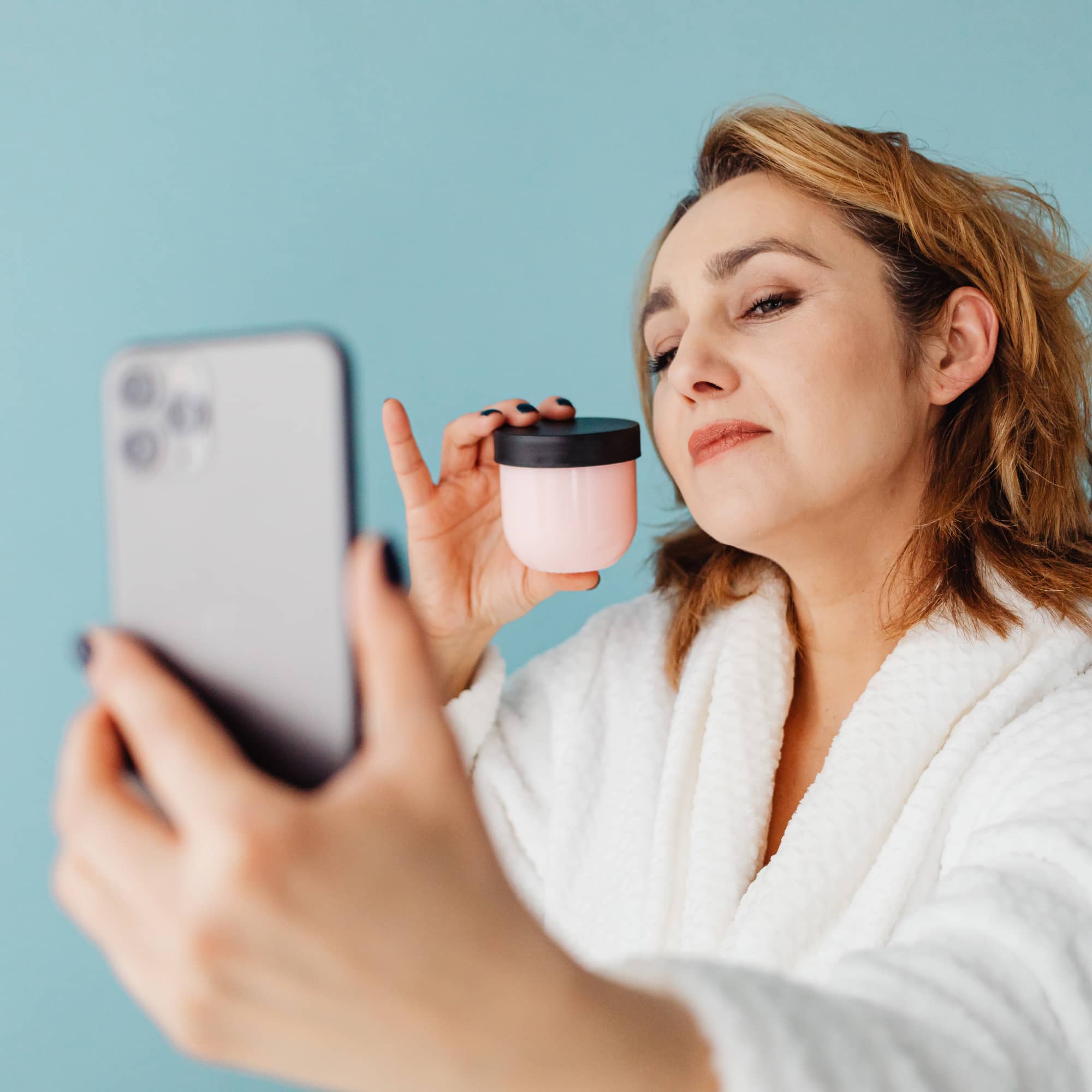 Woman in a bathrobe holding a skincare cream jar while taking a selfie with her smartphone against a blue background.