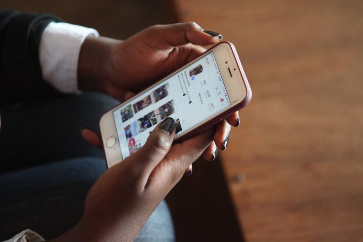 A close-up shot of a person's hands holding a white smartphone. The phone screen displays an Instagram user profile, showing profile statistics, an 'Edit Profile' button, and a grid of photos. The person, who has dark skin and black nail polish, is using their thumbs to interact with the screen. The background consists of a blurred wooden surface.