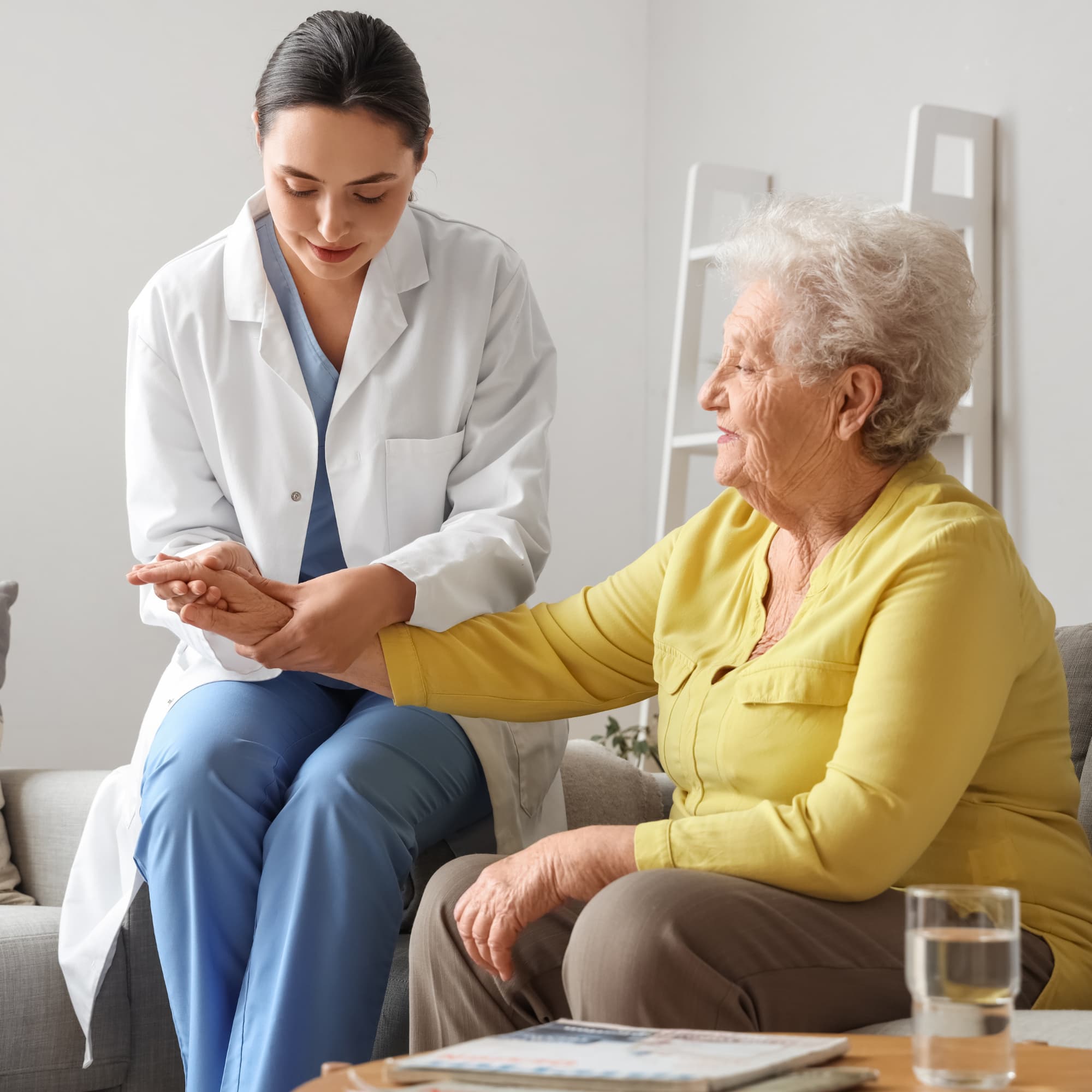 A healthcare professional examining an elderly woman’s hand during a home visit, showing care and medical support in a calm indoor setting.