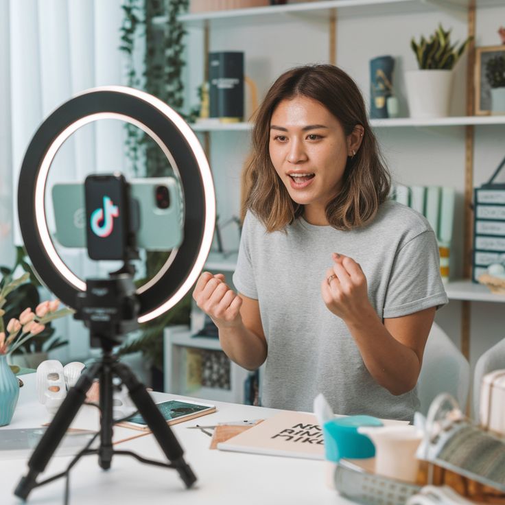 A young woman sits at a white desk, enthusiastically recording a video for TikTok using a smartphone mounted inside a glowing ring light on a tripod. She gestures with her hands while speaking to the camera in a bright, modern room filled with shelves and decorative items.