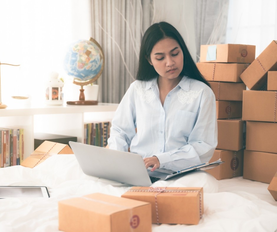 Gemini said
A woman in a light blue shirt sitting on a bed and working on a laptop, surrounded by numerous cardboard shipping boxes and a globe in the background.