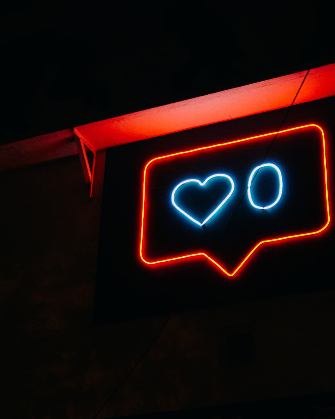 A glowing neon sign against a dark wall resembling an Instagram notification bubble. The bubble is outlined in bright red, containing a blue neon heart icon next to the number zero, also glowing in blue.