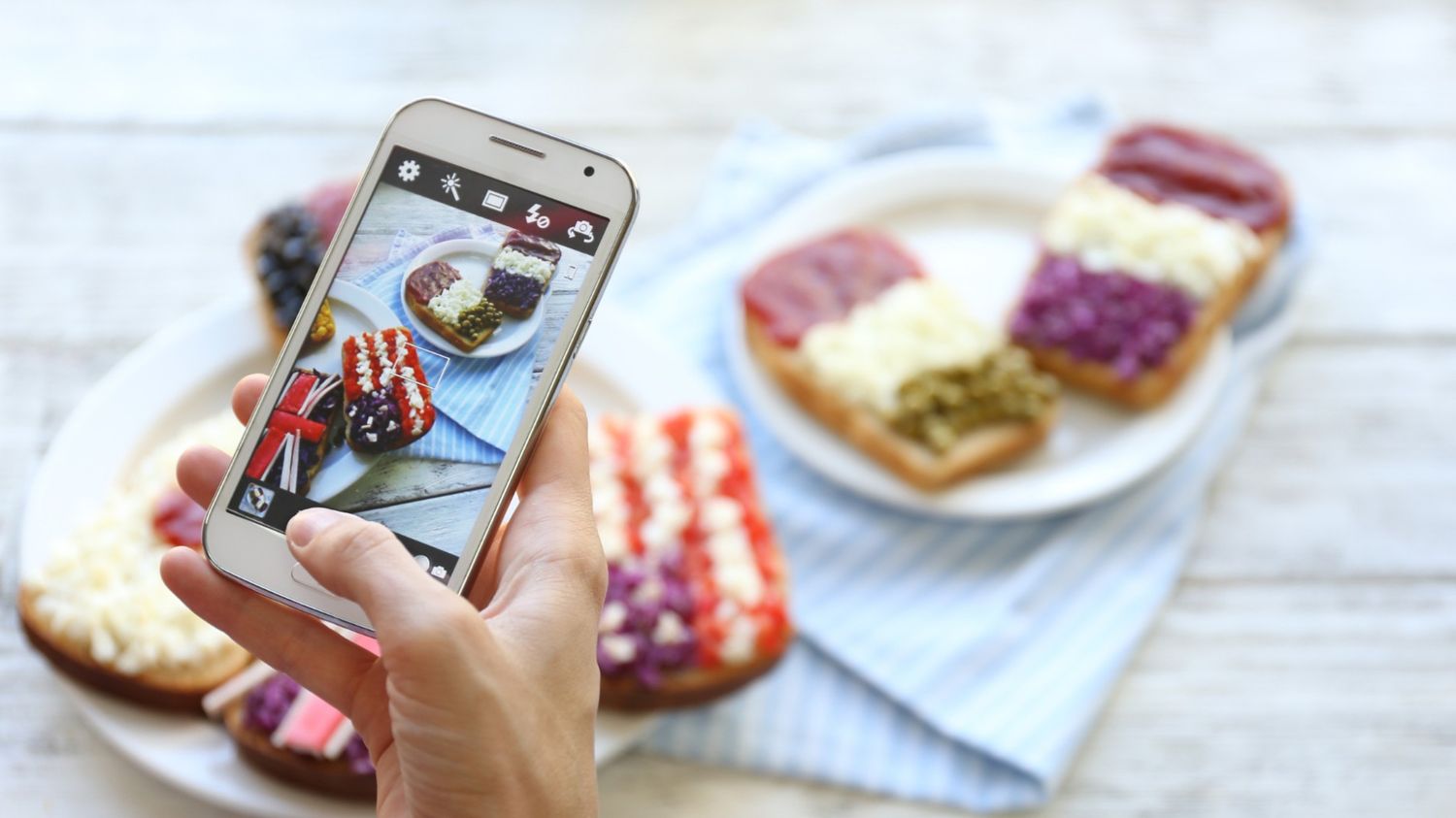 A hand holding a smartphone to take a photo of gourmet toast decorated with colorful toppings like red berries, white cream, and purple cabbage, representing the aesthetic content creation style often seen on Instagram.