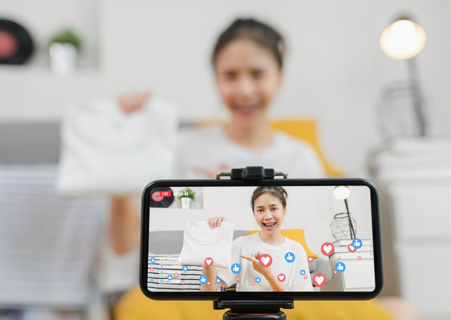 A young woman is hosting a live-streaming session on her smartphone, showcasing a white garment to her audience. The screen is filled with real-time engagement markers, including floating hearts and "like" icons, while a "LIVE" timer in the corner indicates an active broadcast.