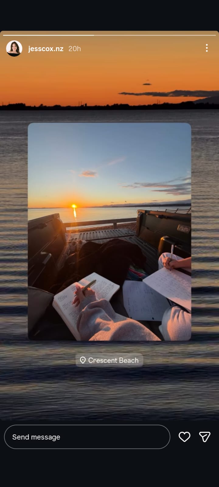 Instagram Story screenshot showing two people writing in notebooks while sitting in the back of a pickup truck parked by the water at sunset. The sun is low on the horizon, reflecting across the calm sea, with an orange and blue sky creating a peaceful backdrop. The story is tagged at Crescent Beach.