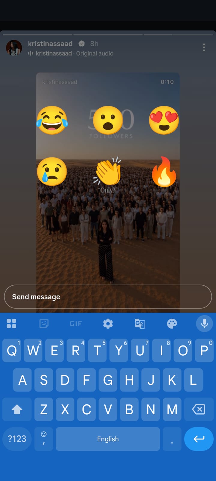 Instagram Story screenshot showing a viral-style post celebrating reaching 500 followers, featuring a person standing in front of a large crowd in a desert landscape. Large reaction emojis (laughing, surprised, heart eyes, worried, clapping, and fire) are overlaid on the video, while the Instagram message field and on-screen keyboard are visible at the bottom, indicating the viewer is preparing to reply to the story.
