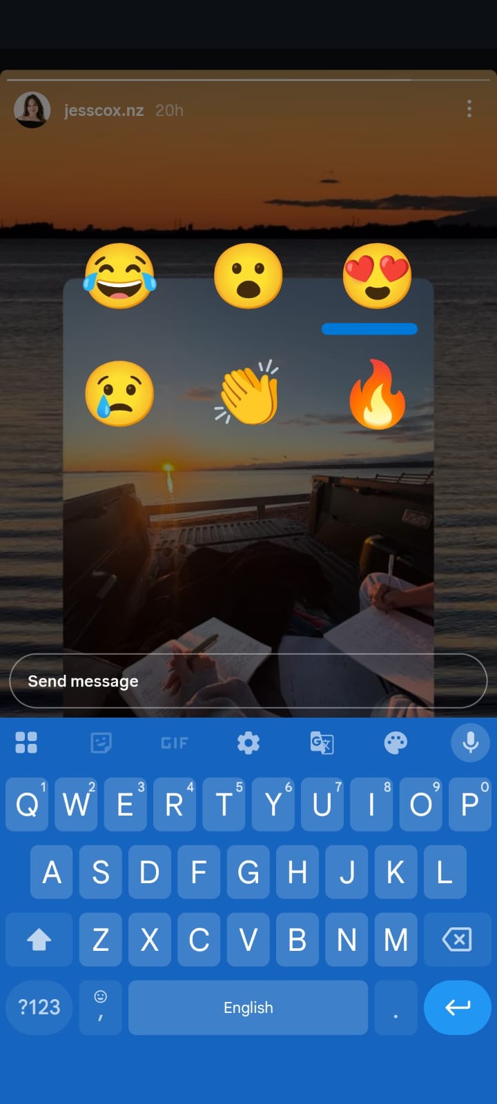 Instagram Story screenshot showing a sunset beach scene viewed from the back of a pickup truck, where two people are writing in notebooks. Large reaction emojis (laughing, surprised, heart eyes, worried, clapping, and fire) are displayed over the story. The Instagram reply field and smartphone keyboard are visible at the bottom, indicating the user is responding to the story.