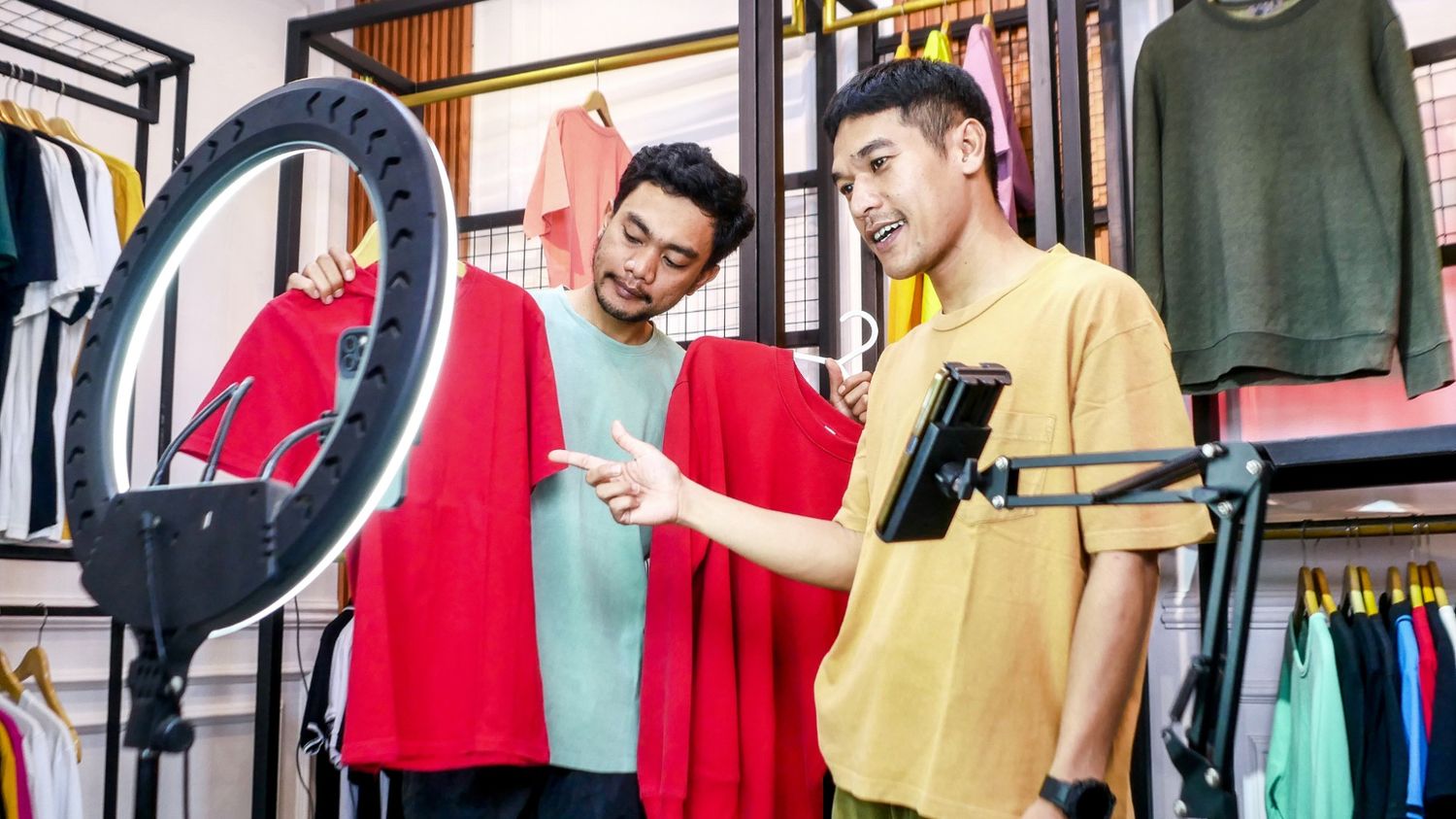 Two men hosting an interactive live stream in a clothing store, using a ring light and smartphone setup to showcase red t-shirts to an online audience.