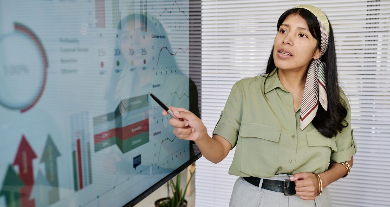 A professional woman in an office setting pointing to a large touchscreen monitor displaying various business analytics, including growth charts, line graphs, and data visualizations, representing interactive marketing strategies and performance tracking.