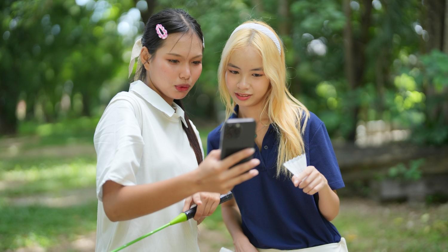 Two young women standing outdoors in a park, pausing their badminton game to look intently together at a smartphone screen. The woman on the left holds the phone and a racket, while the blonde woman on the right holds a shuttlecock.