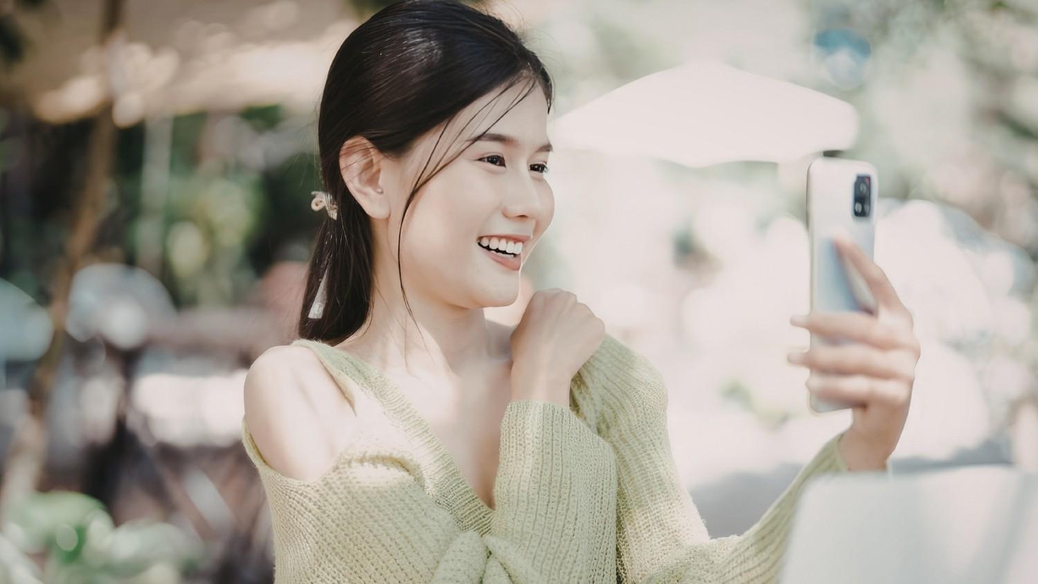 A cheerful young woman outdoors on a bright day, smiling widely as she holds her smartphone up in front of her, appearing to take a selfie or engage in a video call. She is wearing a light green knit top against a softly blurred background.