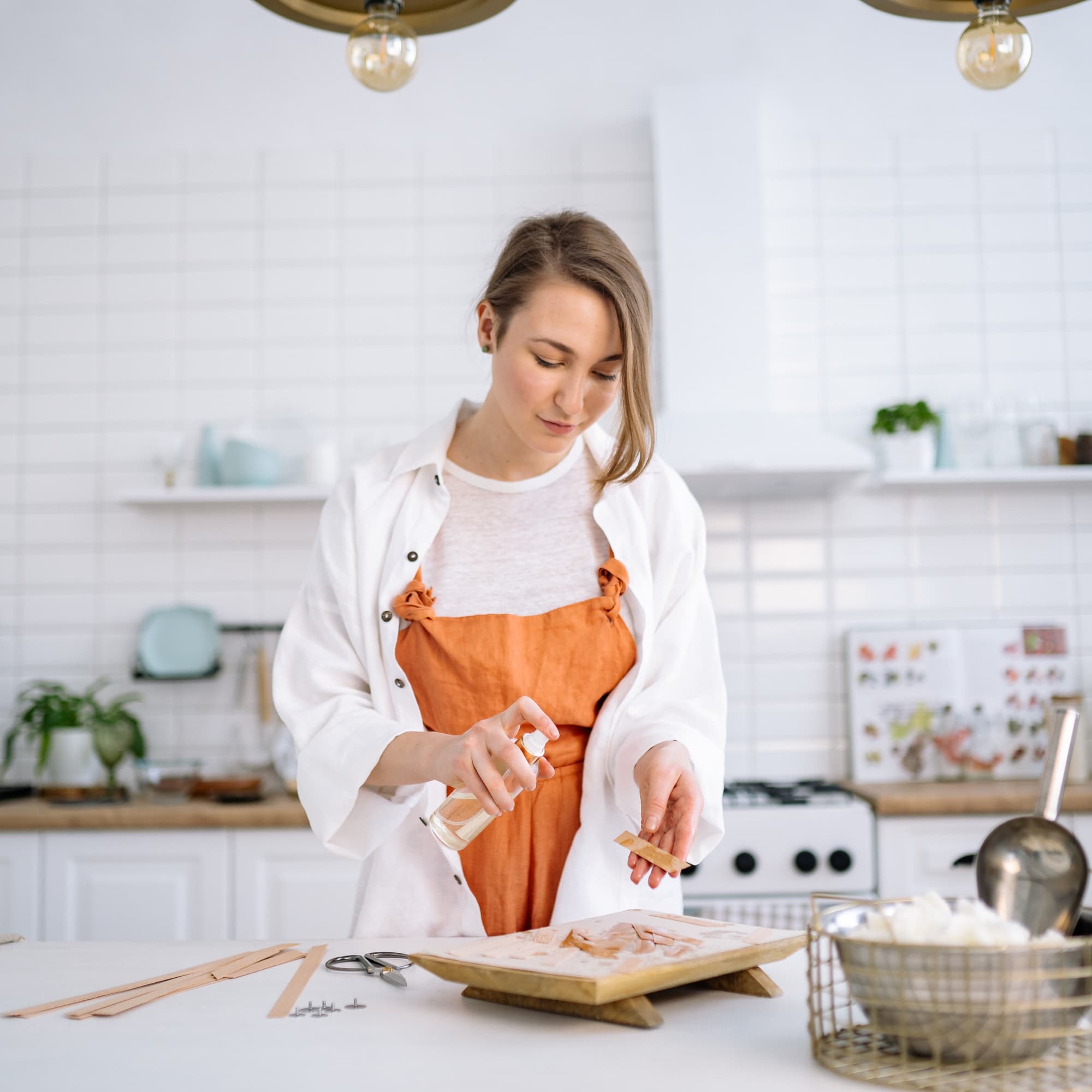 Woman in an orange apron spraying liquid onto wooden sticks on a tray in a bright, modern kitchen.