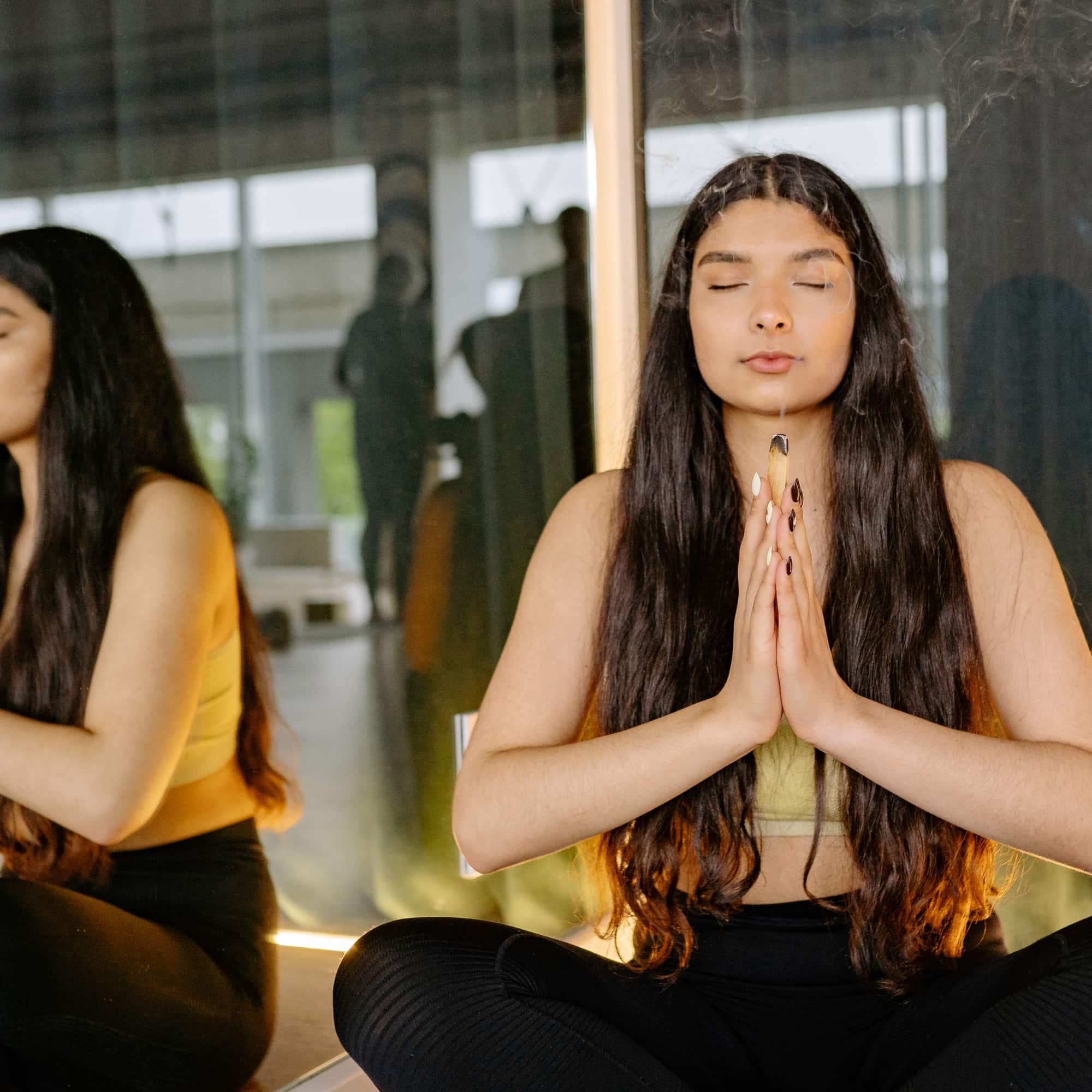 A woman sits cross-legged with eyes closed, hands in a prayer position, meditating indoors beside a mirror with soft warm lighting.