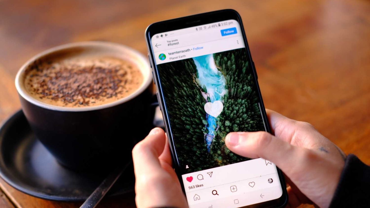 Two hands holding a phone showing an Instagram post of a forest river, beside a cappuccino on a wooden table