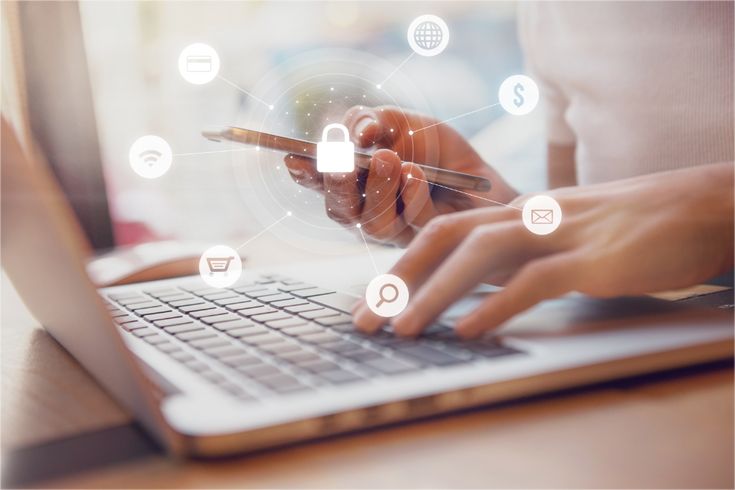 A close-up of a person's hands simultaneously holding a smartphone and typing on a laptop keyboard. Overlaid on the image is a glowing digital network graphic featuring a central white padlock icon. Thin white lines connect the padlock to surrounding circular icons representing a credit card, a globe, a dollar sign, an envelope, a magnifying glass, a shopping cart, and a Wi-Fi symbol.