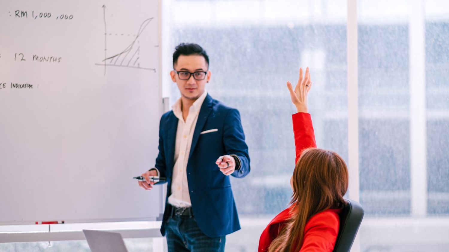 This image features a presenter in a navy blazer pointing toward an audience member in a bright red jacket who has her hand raised. In the background, a whiteboard displays a growth chart and the text "12 MONTHS," indicating a long-term strategic discussion.  Continuing with your professional narrative on Instagram and lead generation, this visual captures the interactive phase of lead qualification.