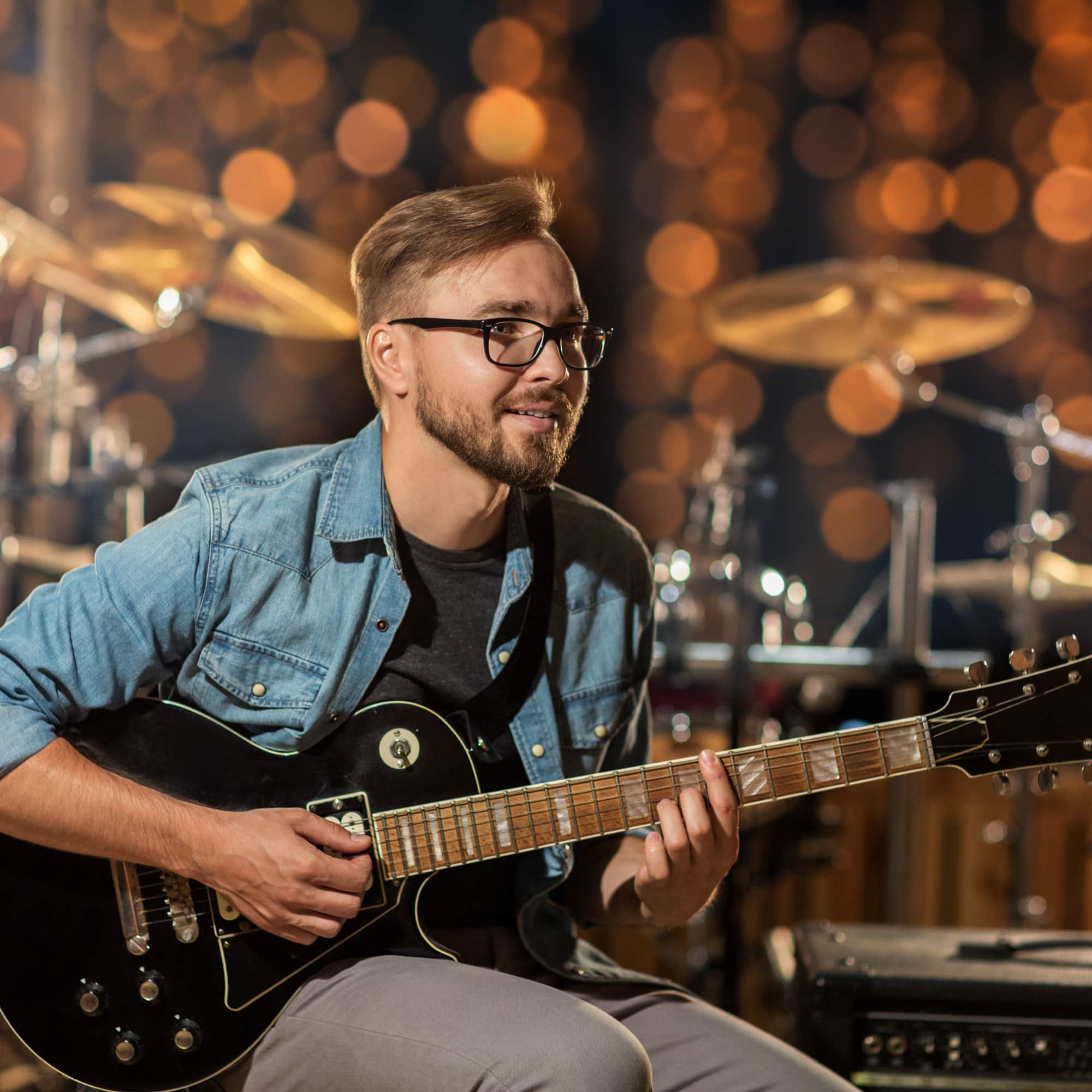 Man playing an electric guitar on stage, smiling during a live performance with drums and warm bokeh lights in the background.